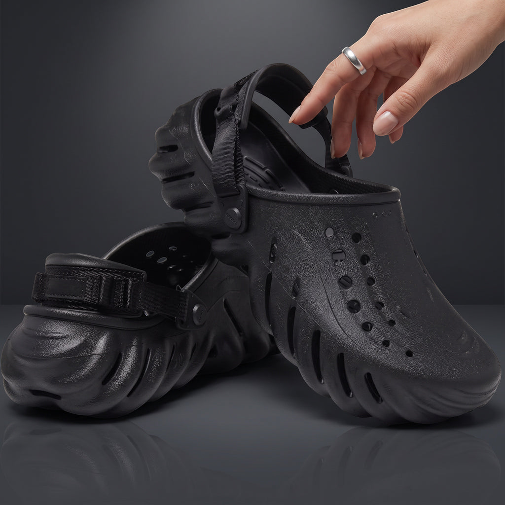 Sabots Echo Clog noirs LAS ROSAS en mousse sculptée avec bride réglable, tenus par une main sur fond gris.